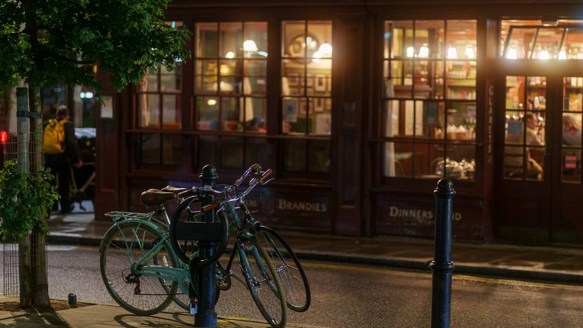 Two bicycles locked to a stand outside a London coffee house at night, its windows lit warmly from inside