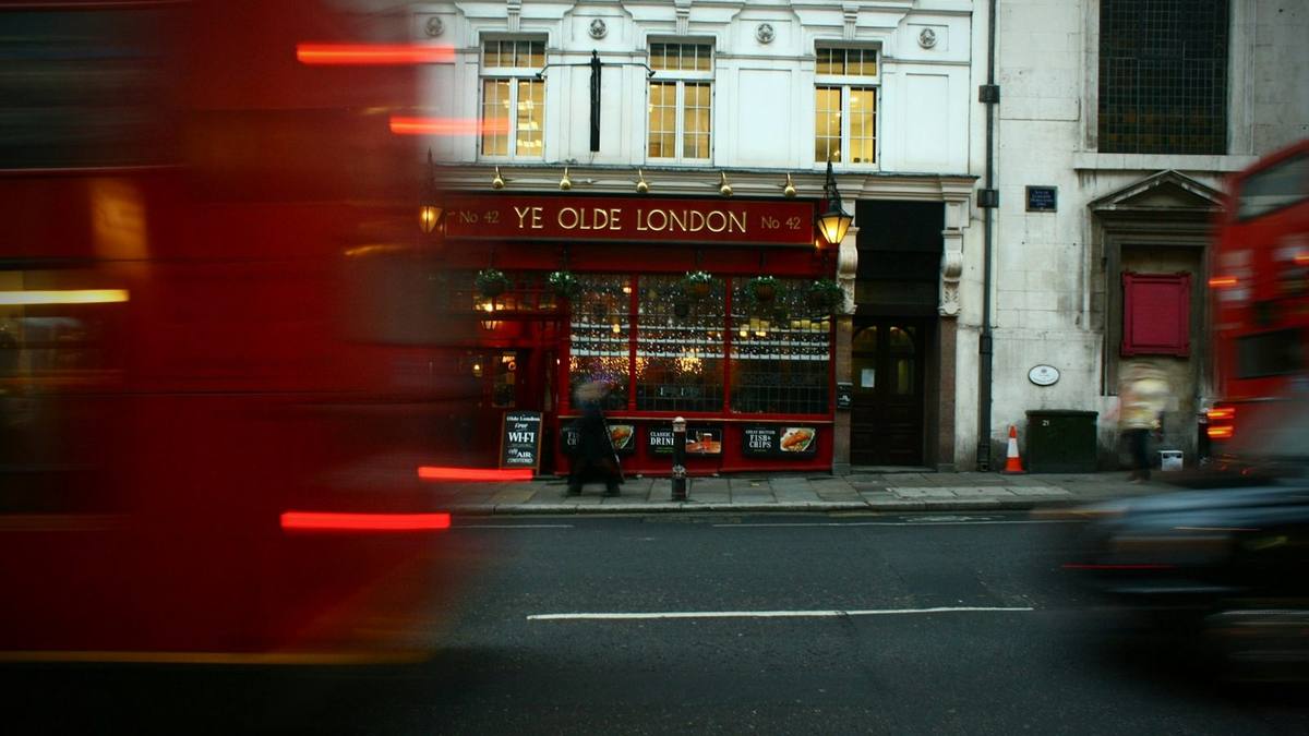 A red London bus blurs past a traditional pub on a City of London street, with a taxi passing on the right