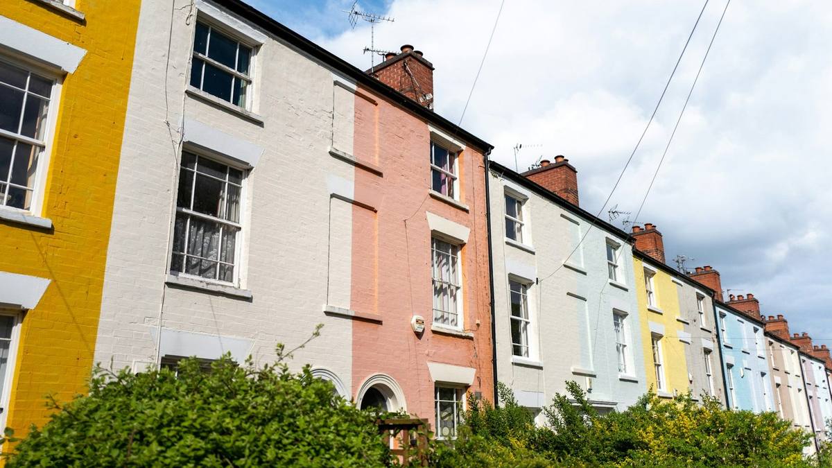 A row of three-storey terraced houses painted yellow, white, pink and pale blue, with sash windows and green hedges in front, under a blue sky