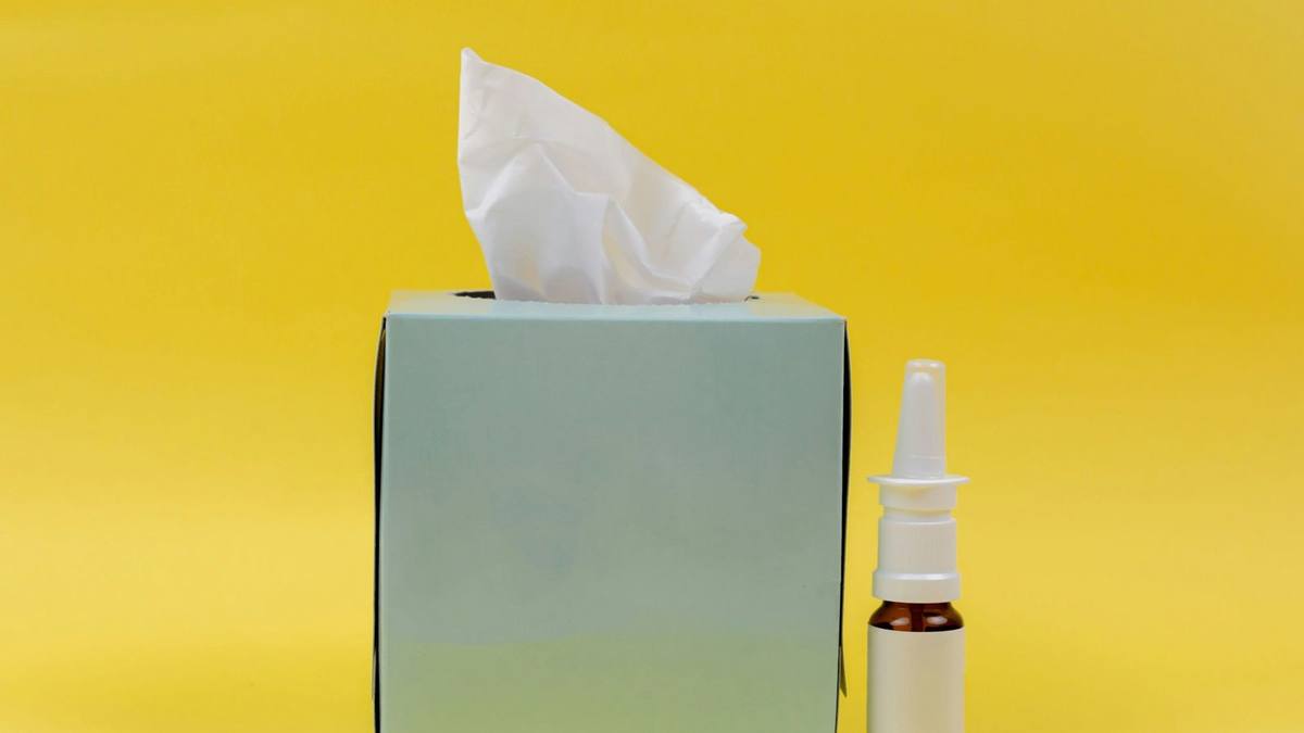 A box of tissues and a bottle of nasal spray against a plain yellow background