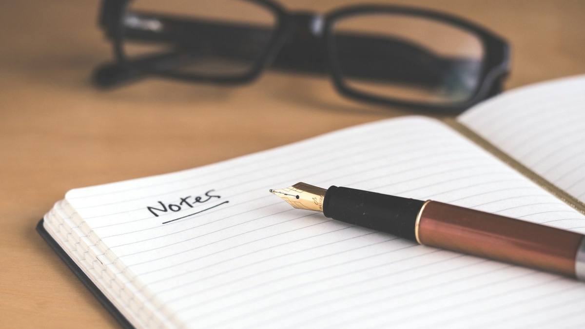 A fountain pen resting on an open lined notebook headed Notes, with reading glasses behind