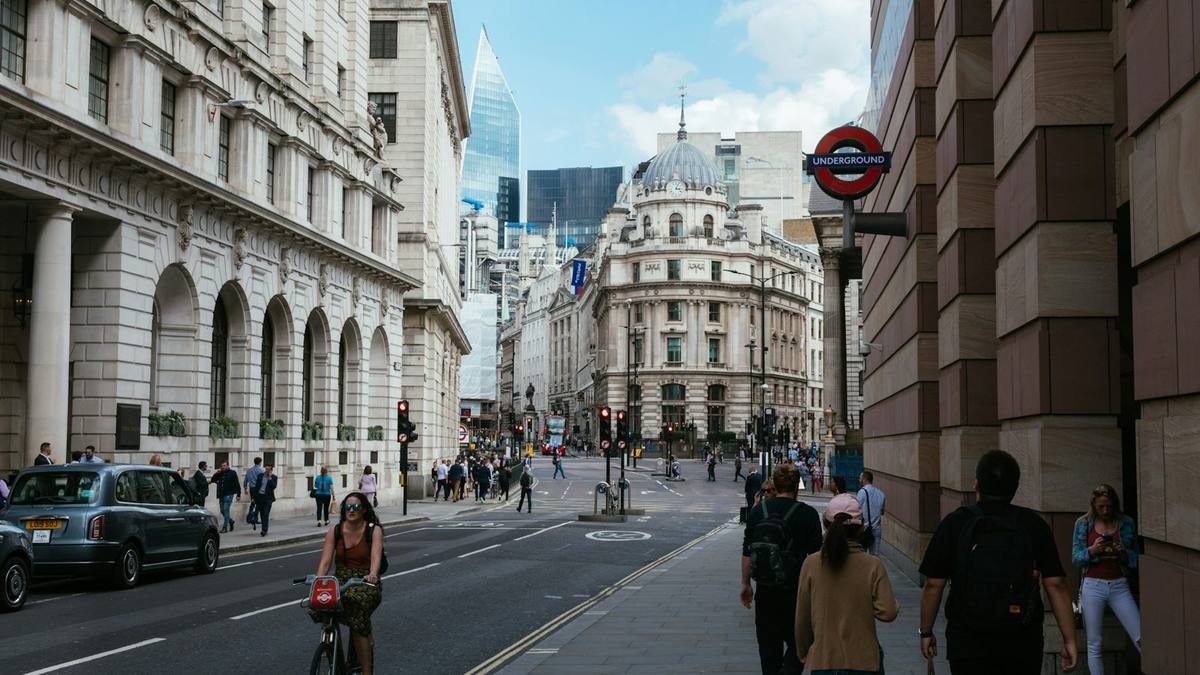 People walking and cycling along a street of stone office buildings in the City of London, with an Underground sign on the right