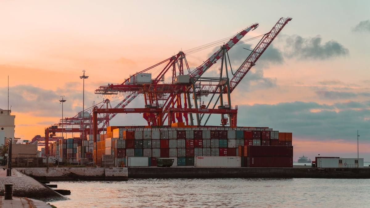 Red container cranes and stacked shipping containers at a port under a sunset sky