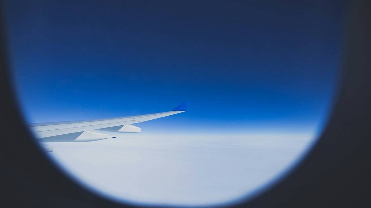 View through an aircraft window of a white wing with a blue wingtip above a flat layer of white cloud