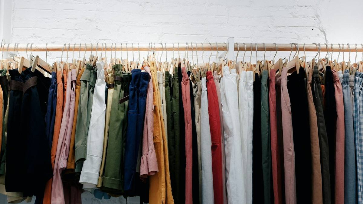 A long rail of trousers and shirts in assorted colours hanging on wooden hangers against a white brick wall