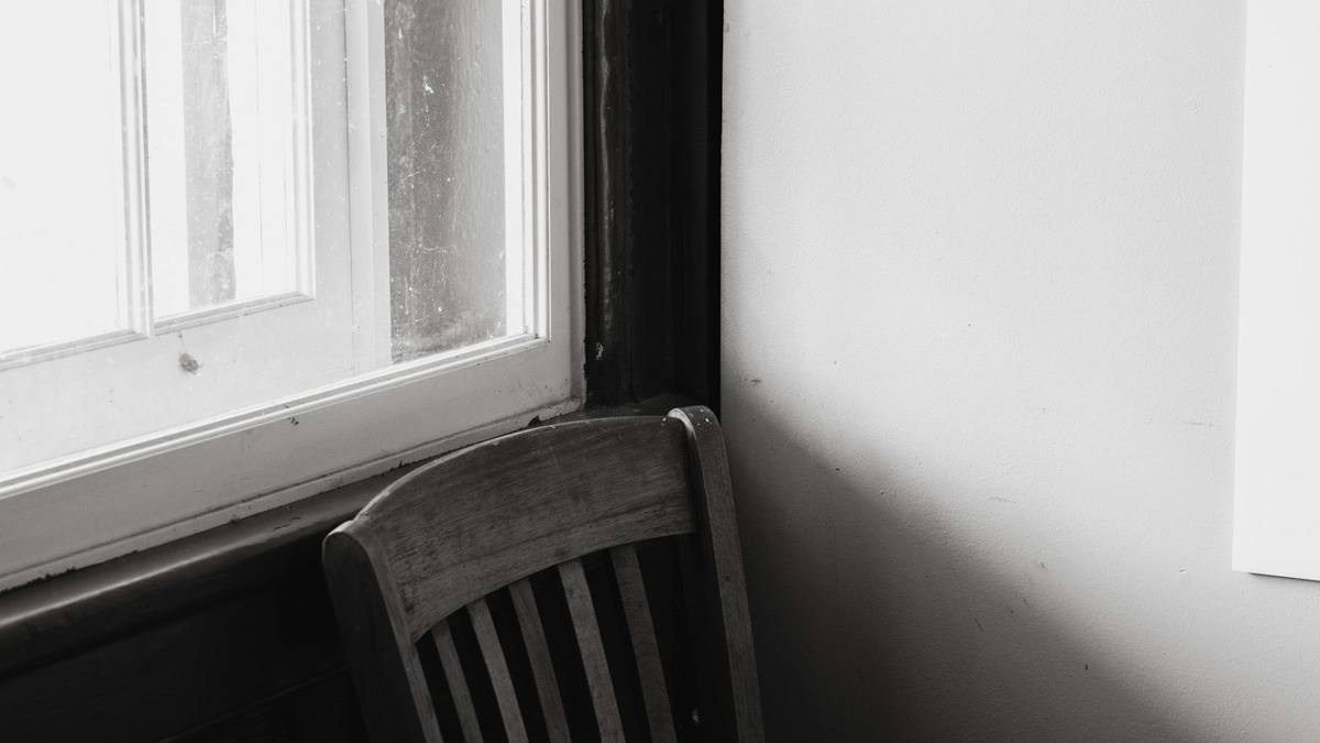 An empty wooden chair beside a bright sash window and a worn wooden table